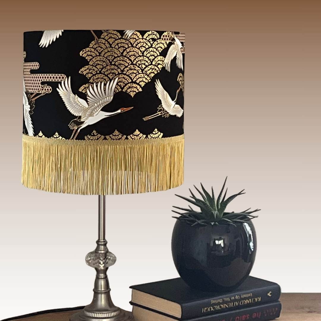 Art Deco style Japanese Cranes Lampshade that features cranes in flight and Art Deco-style clouds on a black background with gold metallic highlights and fringe on a table