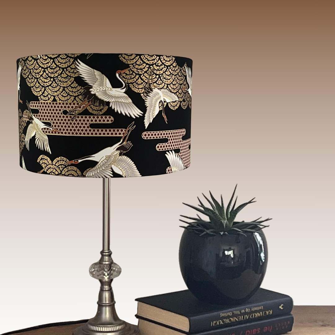 Art Deco style Japanese Cranes Lampshade that features cranes in flight and Art Deco-style clouds on a black background with gold metallic highlights styled on table.