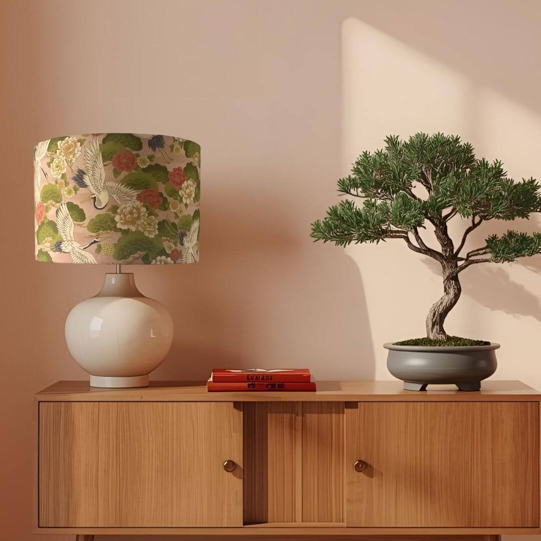Decorative lampshade with Japanese cranes and florals pattern on a peachy pink background on a sideboard with a bonsai tree