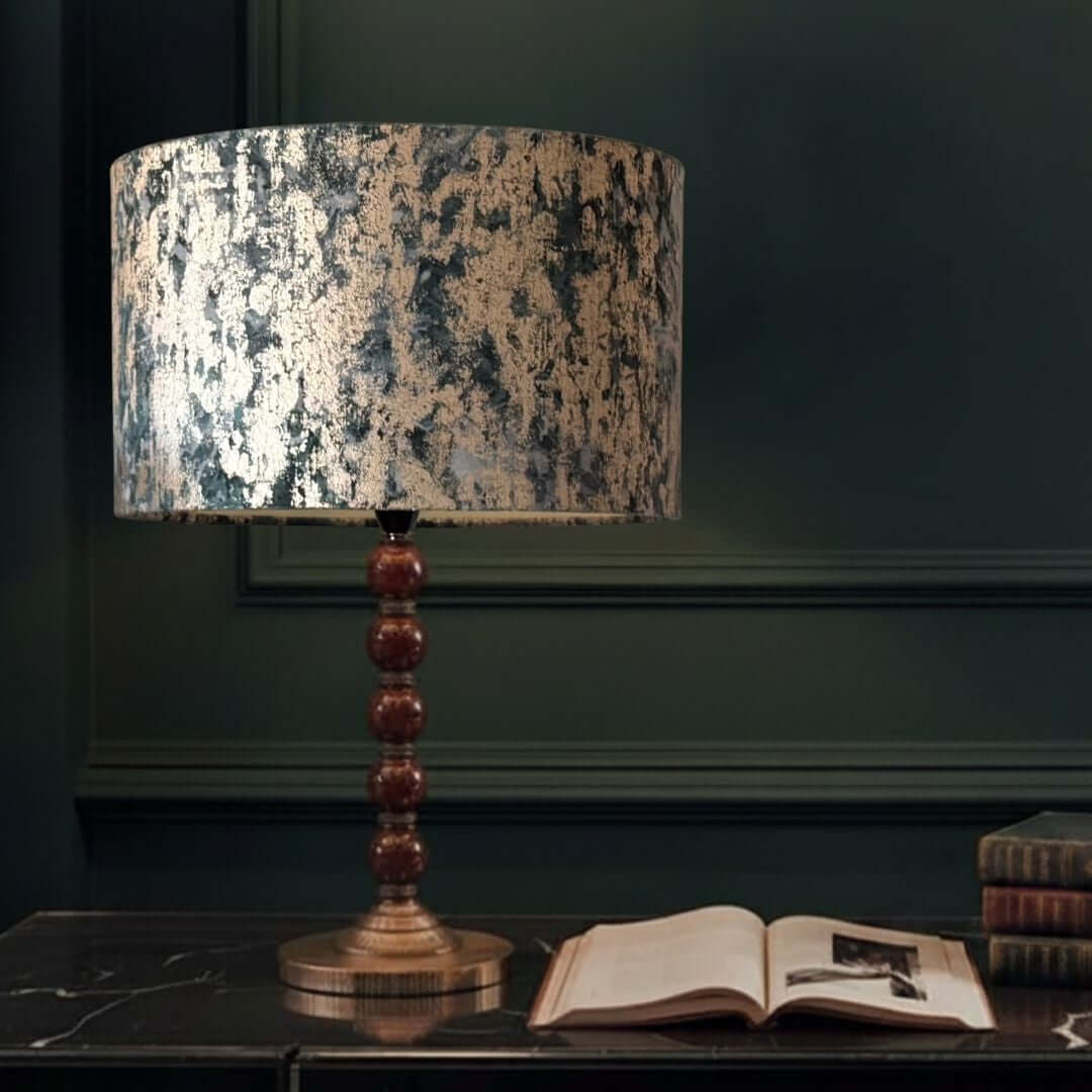 Clarke & Clarke Dipinto velvet lampshade in the teal colourway shown on a table with books on a green panelled background.