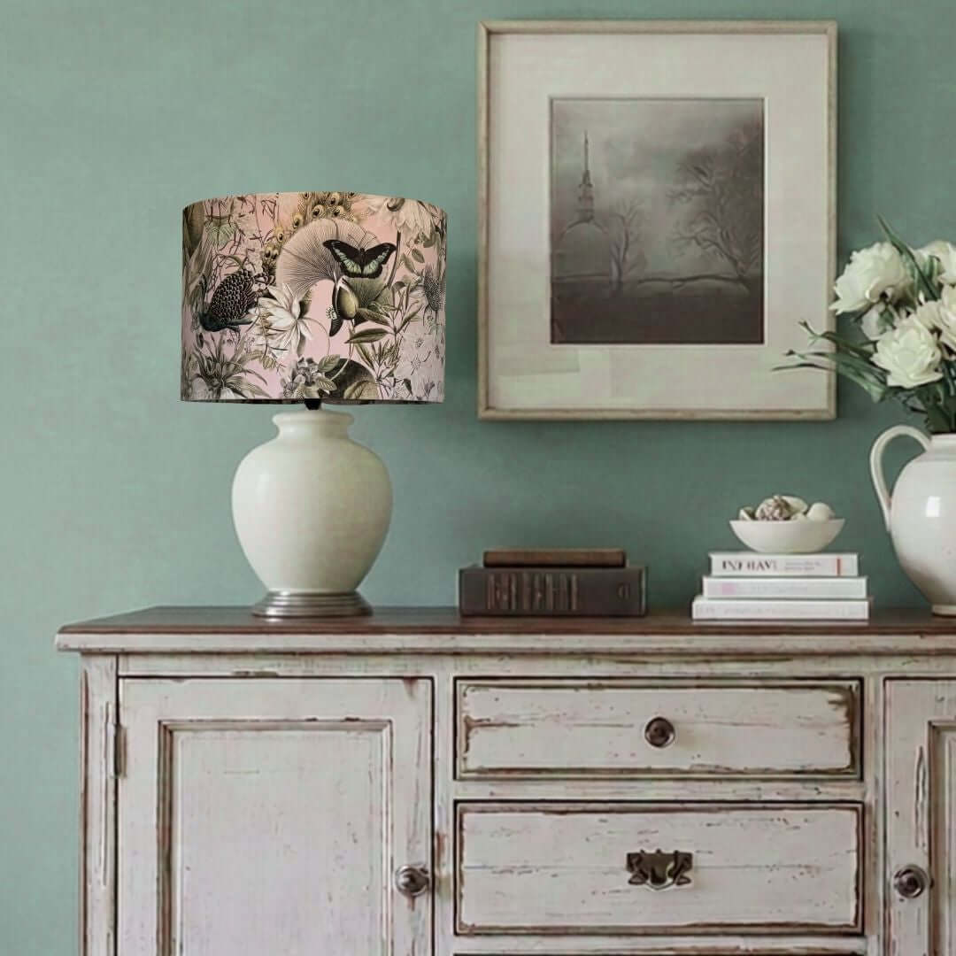 A table lamp with a light-coloured ceramic base and a velvet drum lampshade featuring a peacock, butterfly, and floral print in muted pink, green, and teal tones, styled on a distressed white wooden sideboard against a sage-green wall.