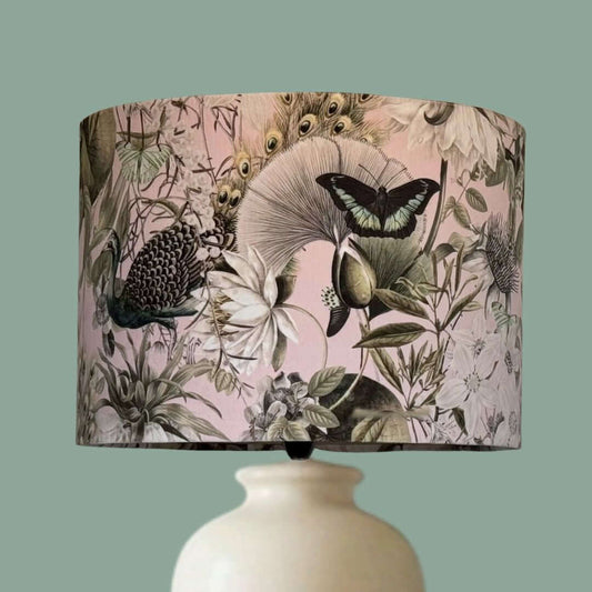 Velvet drum lampshade with a detailed botanical print with peacocks, butterflies and florals in pale pink, white, sage green and teal. Shown on a ceramic lampbase.