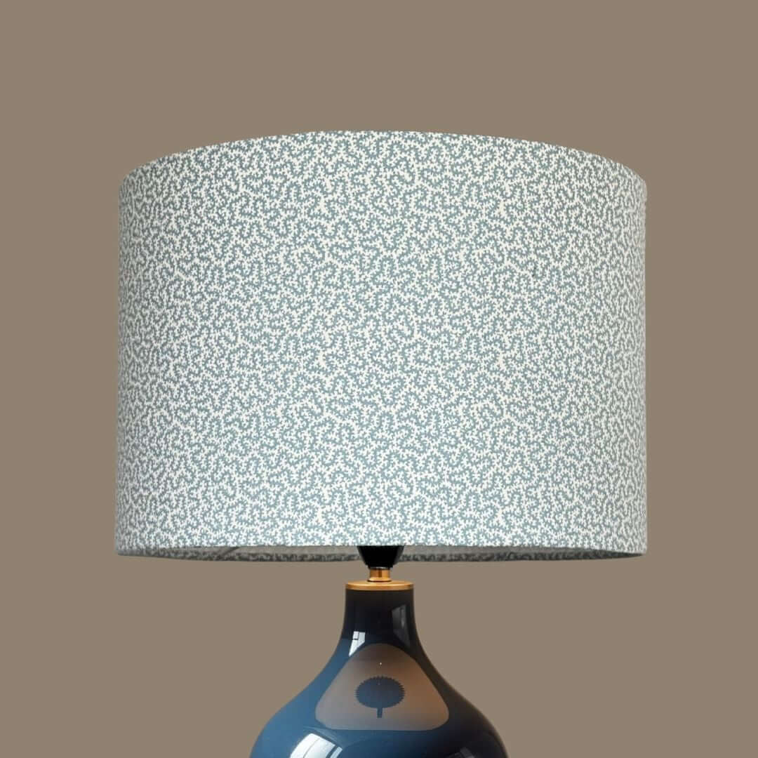 A table lamp with a drum lampshade crafted from Ian Sanderson Petra fabric in Chalk Blue. A small-scale abstract pattern of light blue and white, resting on a glossy dark blue ceramic base.