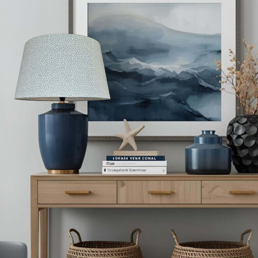 Empire conical lampshade in Ian Sanderson Petra fabric in Chalk Blue shown in decorative setup with a lamp, books, and decorative items on a wooden console table.