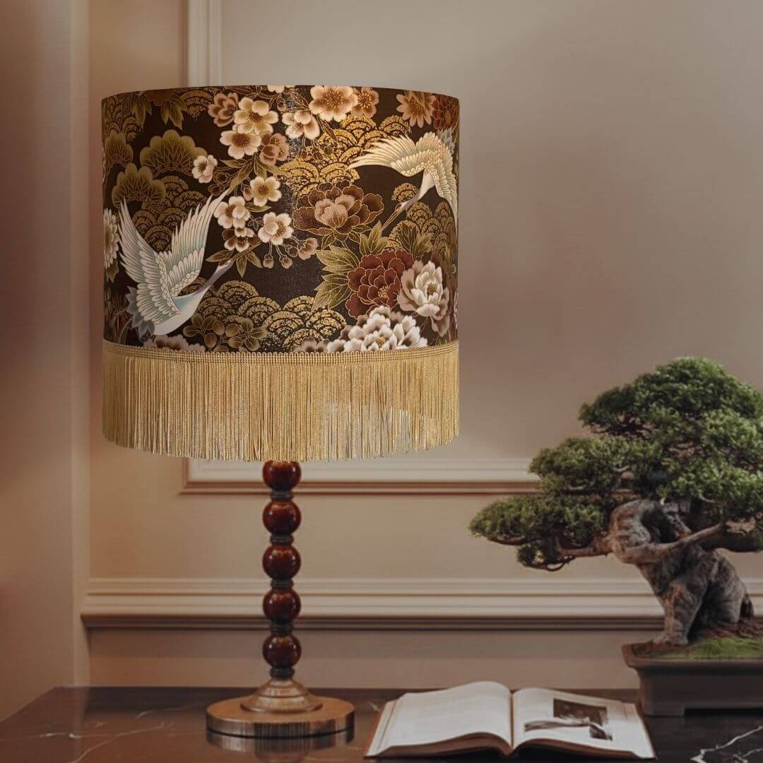 A decorative table lamp shade with Japanese cranes, golden clouds, bronze and white peonies and a gold fringe. Styled on a tabletop next to an open book and a bonsai tree.