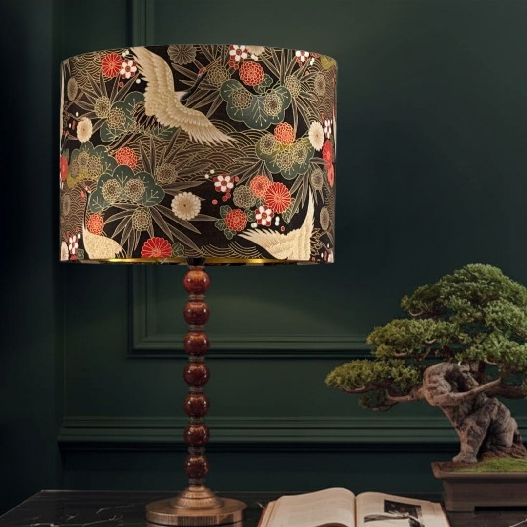 Drum Lampshade made with Authetic Japanese fabric with cranes, florals and metalic gold accents shown styled on a table with a bobble lampsbase and a bonsai tree
