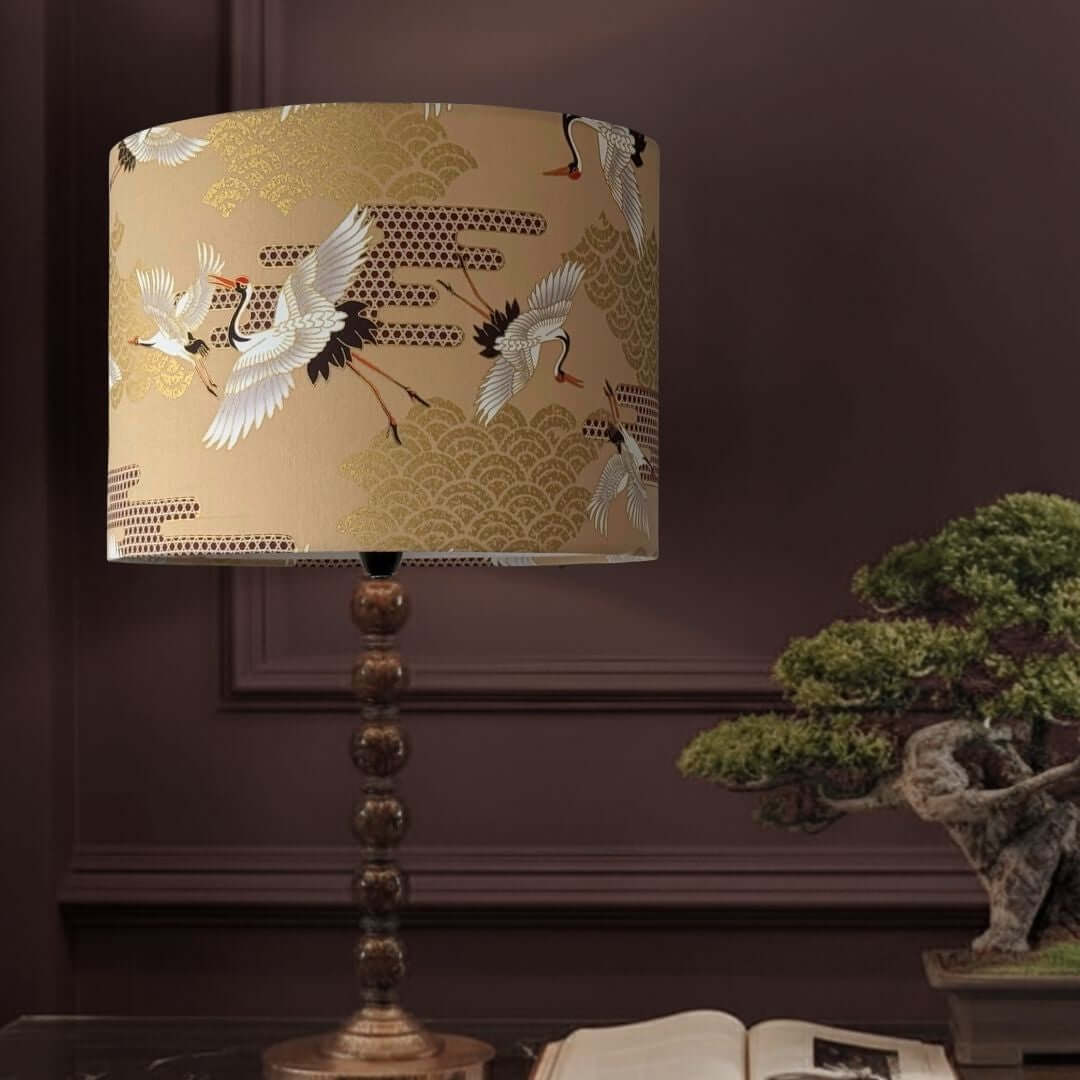 A table lamp with a beige drum shade featuring a printed pattern of white cranes and gold wave motifs, mounted on a dark bronze-toned turned wood pedestal. The lamp is styled in a room setting against a dark brown paneled wall, with a bonsai tree and an open book in the background.