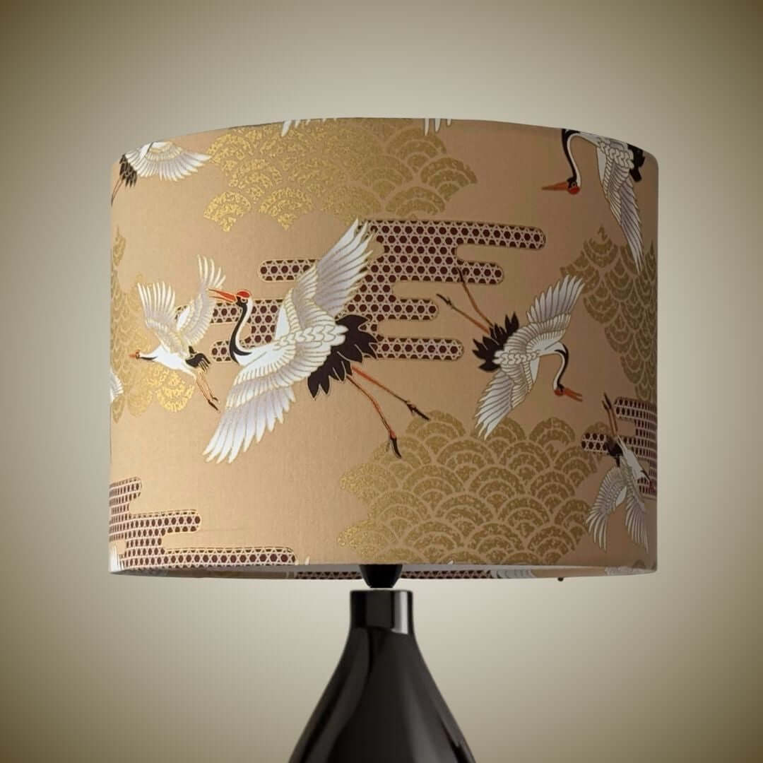 A table lamp with a beige drum shade featuring a pattern of white cranes and gold wave motifs, mounted on a black ceramic base.