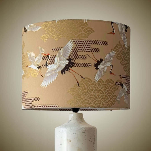 A table lamp with a beige drum shade featuring a pattern of white cranes and gold wave motifs, mounted on a textured white ceramic base.