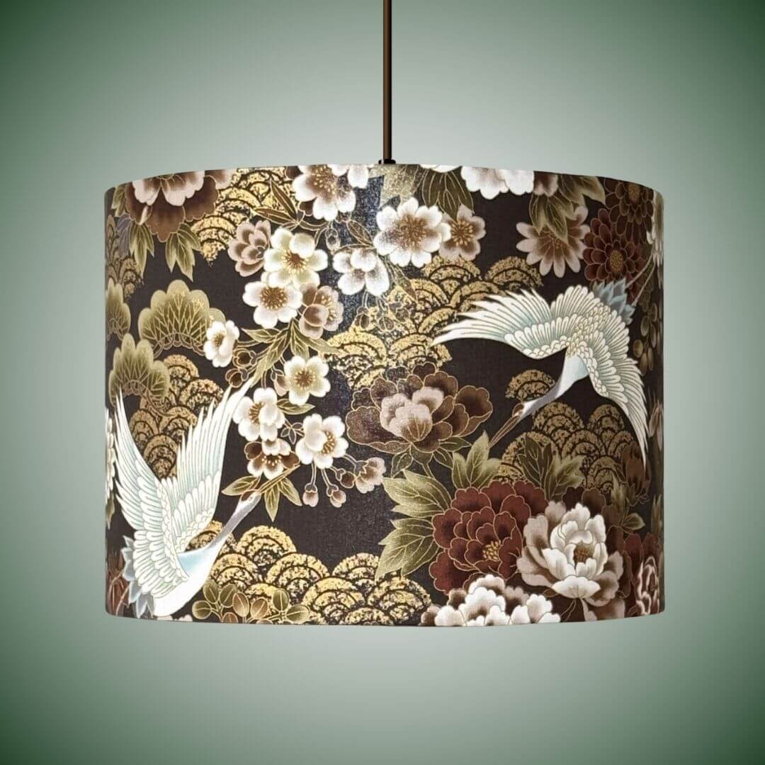 Japanese cranes and florals drum lampshade with metallic accents and a dark brown background.  With an intricate Japanese-style print featuring white cranes, white and bronze peonies and gold wave patterns. Shown on a ceiling pendant.