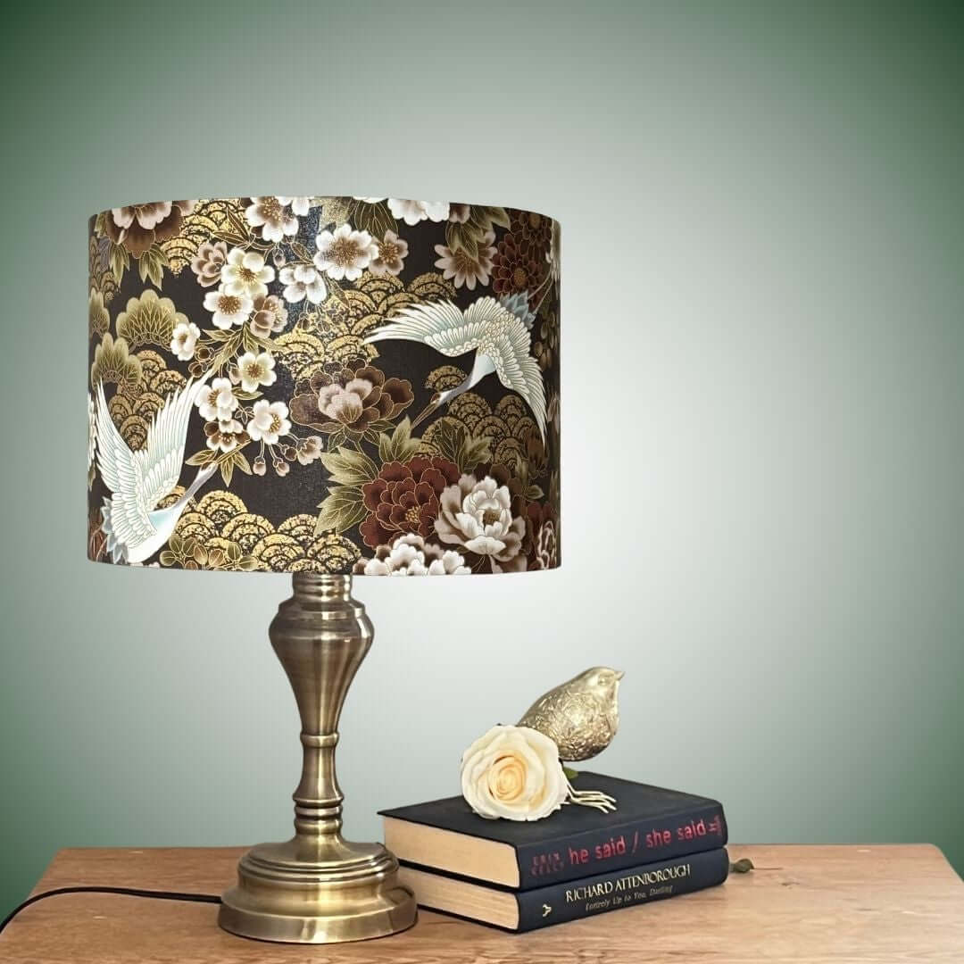 Japanese cranes and florals drum lampshade with metallic accents shown on a brass lampbase on a table