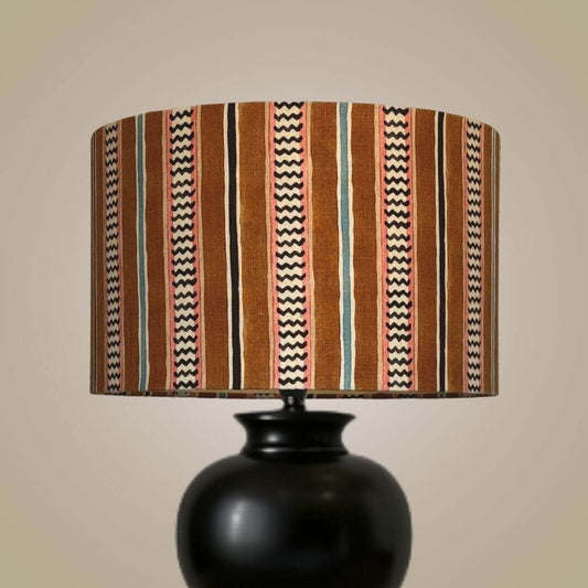 A table lamp with a black ceramic base and a drum lampshade in Linwood hire wire fabric in the Allsorts colourway. Featuring an alternating chevron and solid stripe pattern.