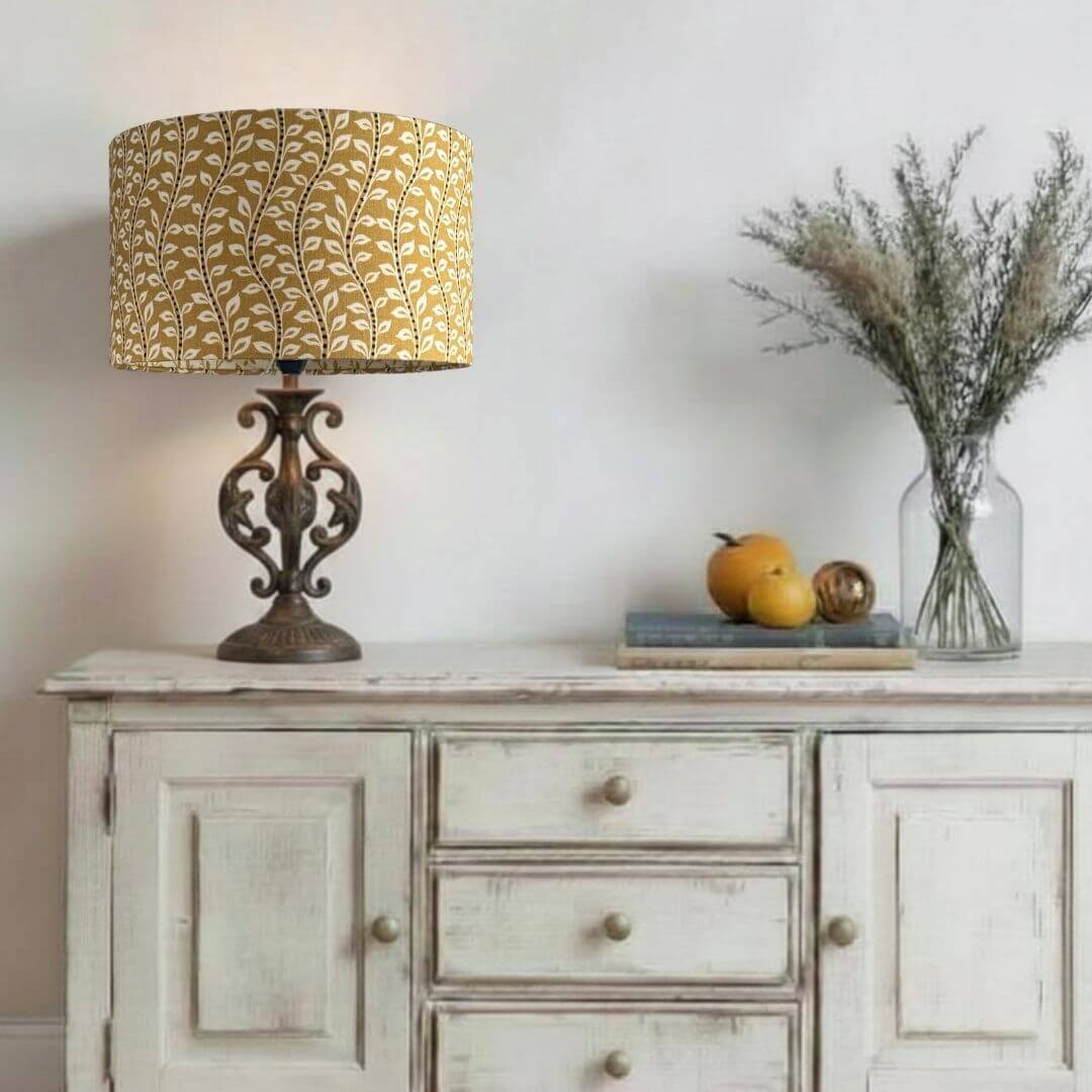 Decorative lamp with a yellow patterned shade on a wooden sideboard with fruit and flowers.