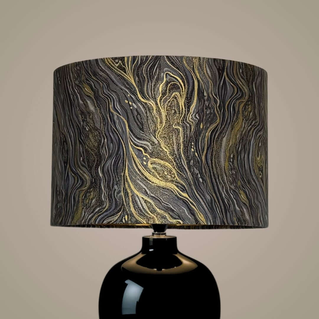 A table lamp with a glossy black ceramic base and a drum shade featuring a silver, grey, and gold marbled pattern.