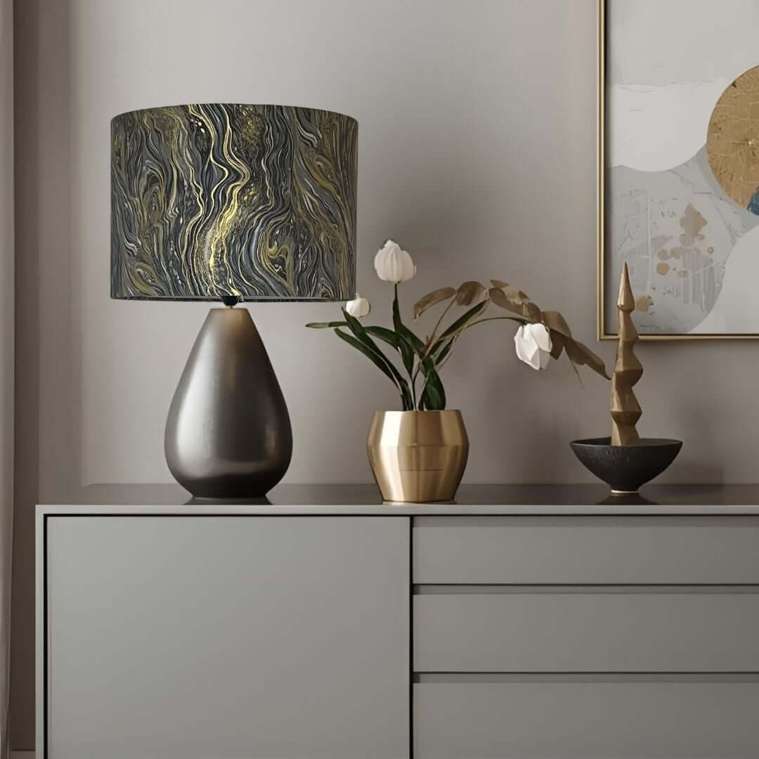 A table lamp with a dark gray teardrop-shaped base and a cylindrical drum shade featuring a black, gray, and gold marbled swirl pattern, styled on a gray sideboard next to a gold vase with tulips.