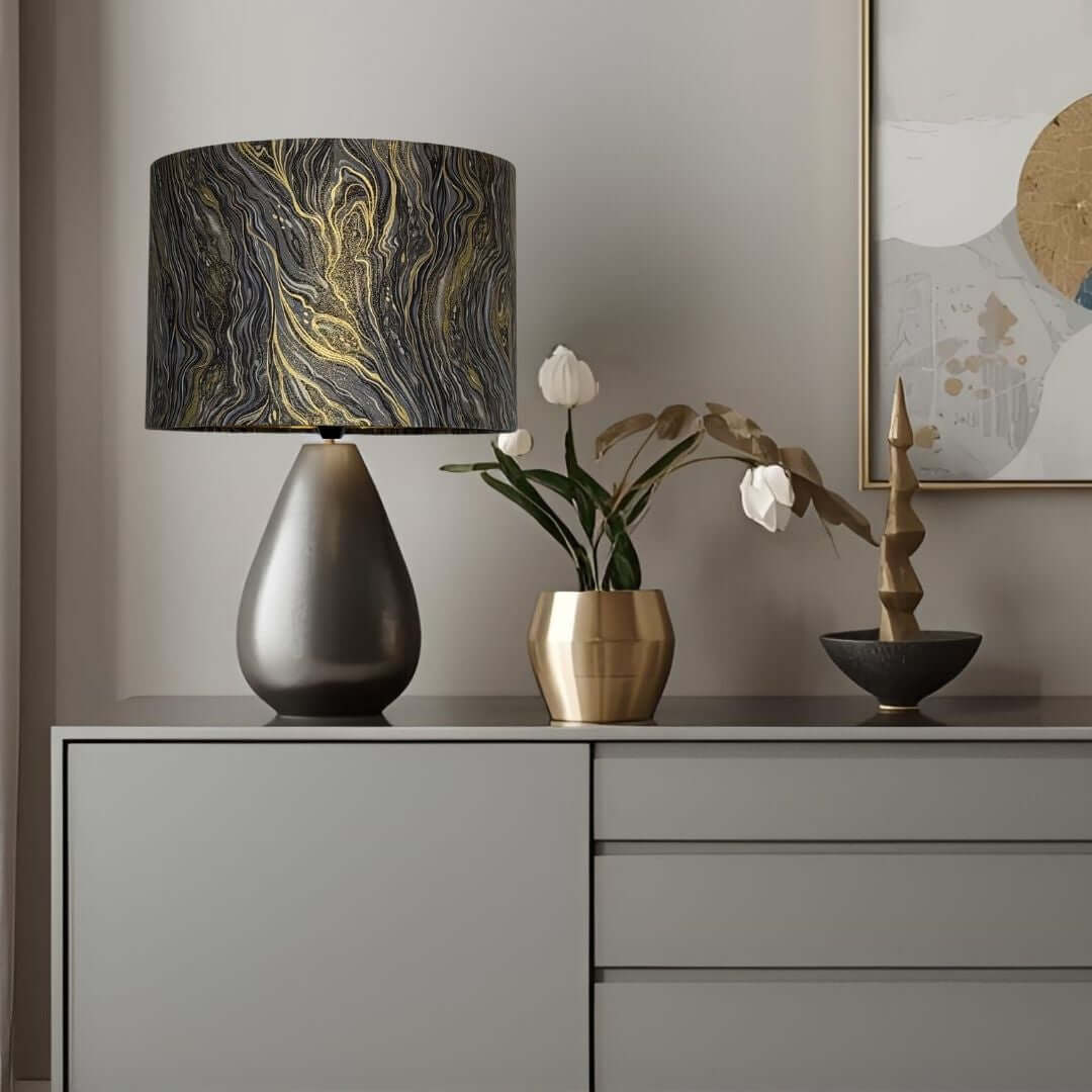 A table lamp with a dark grey teardrop-shaped base and a cylindrical drum shade featuring a silver, black, grey, and gold marbled swirl pattern, styled on a grey sideboard next to a gold vase with tulips.