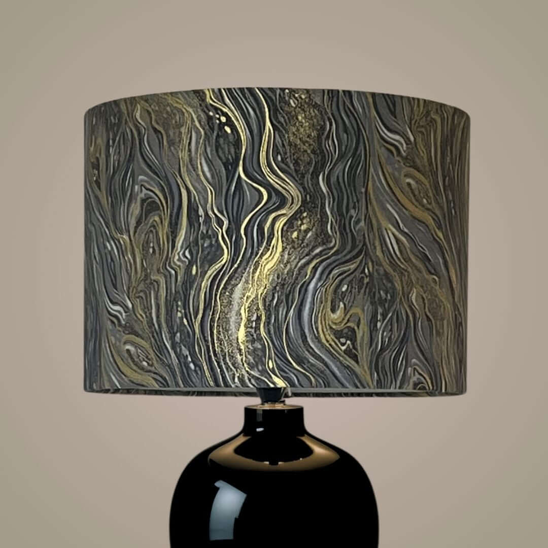 A table lamp with a glossy black ceramic base and a cylindrical drum shade featuring a marbled abstract pattern in black, gray, white, and metallic gold.