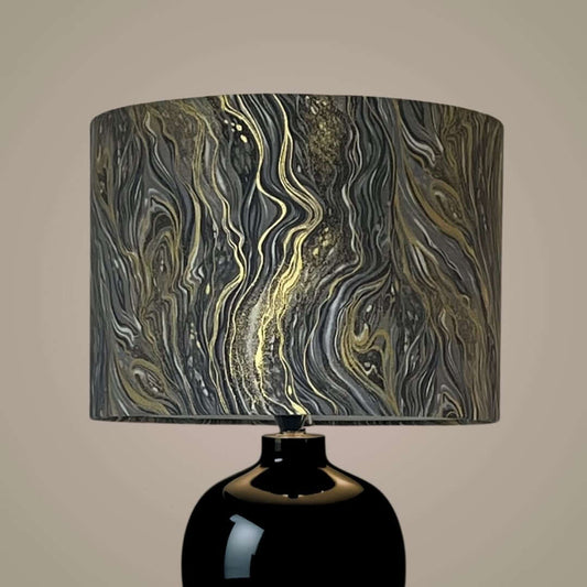 A table lamp with a glossy black ceramic base and a cylindrical drum shade featuring a marbled abstract pattern in black, gray, white, and metallic gold.
