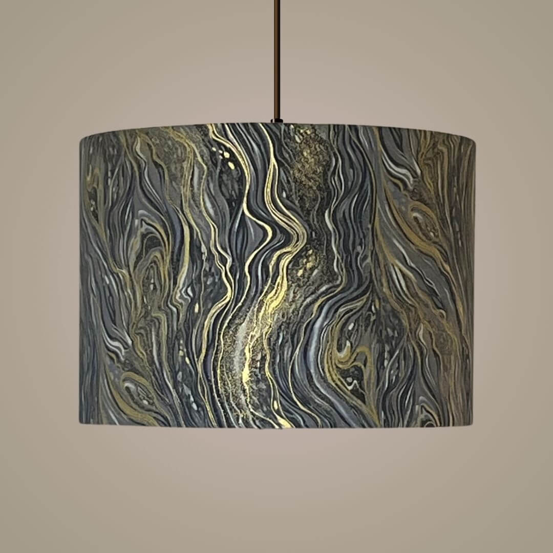 A drum pendant light shade featuring an abstract marbled pattern in black, grey, white, and metallic gold, on a ceiling light