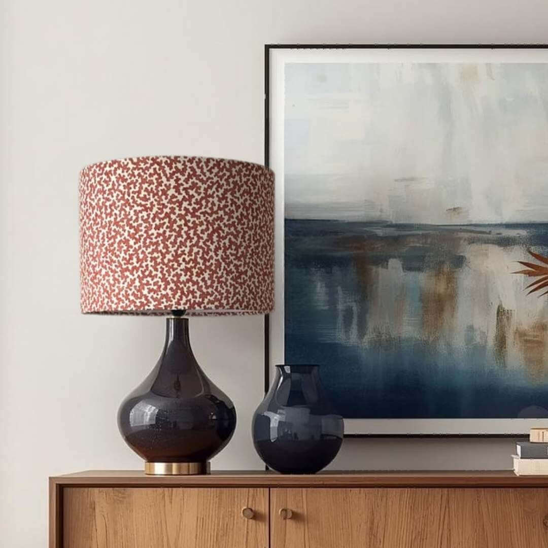 Romo Junie Pimento drum lampshade  on navy lamp base shown on a wooden cabinet with an abstract painting in the background