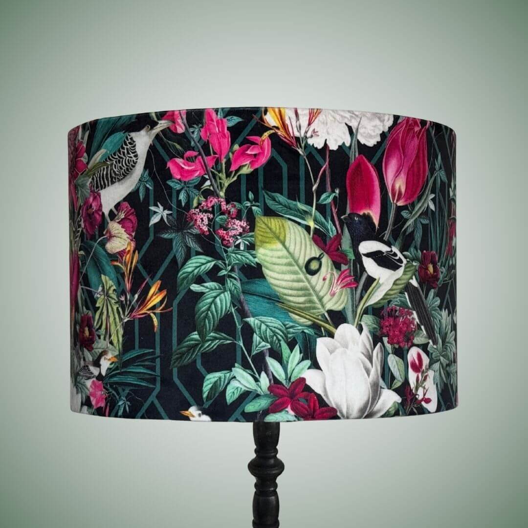 A velvet drum lampshade with a vibrant tropical floral and bird pattern on a dark background, mounted on a black lamp stand 