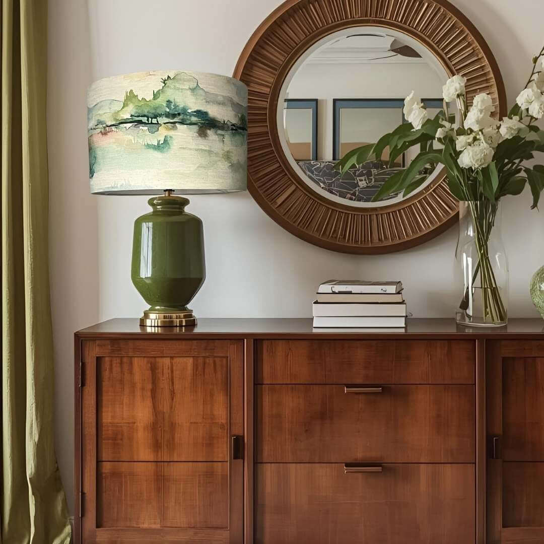 Wooden sideboard with a voyage maison russet shores lampshade on a green lamp base, books, and flowers in a room.