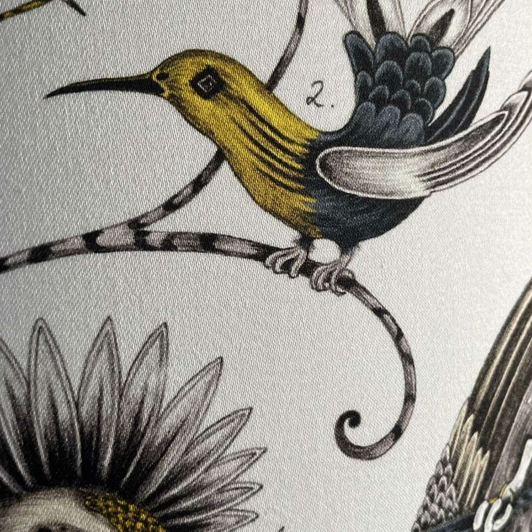 Emma J Shipley Audubon Drum Lampshade featuring exotic birds and foliage in shades of gold, yellow, beige, and charcoal grey on a white background, showing detail of bird