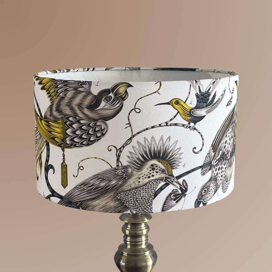 Emma J Shipley Audubon Drum Lampshade featuring exotic birds and foliage in shades of gold, yellow, beige, and charcoal grey on a white background