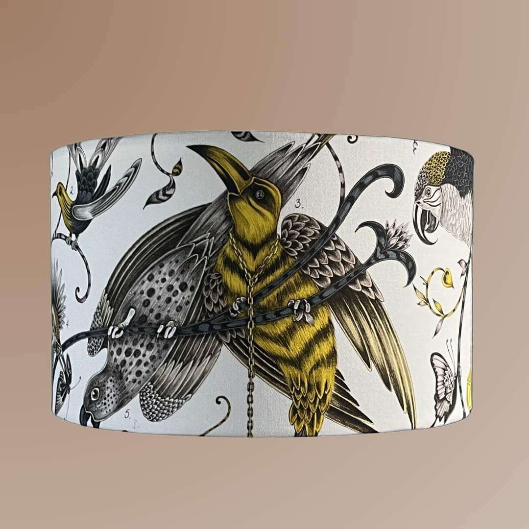 Emma J Shipley Audubon Drum Lampshade featuring exotic birds and foliage in shades of gold, yellow, beige, and charcoal grey on a white background