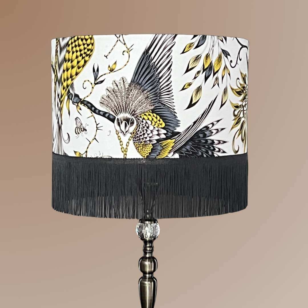 Striking statement lampshade in luxury cotton satin fabric designed by Emma J Shipley. Featuring exotic birds and foliage in shades of gold, yellow, beige, and charcoal grey on a white background, finished with a dark grey fringe