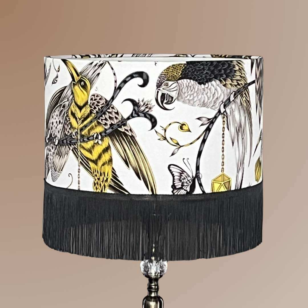 Striking statement lampshade in luxury cotton satin fabric designed by Emma J Shipley. Featuring exotic birds and foliage in shades of gold, yellow, beige, and charcoal grey on a white background, finished with a dark grey fringe