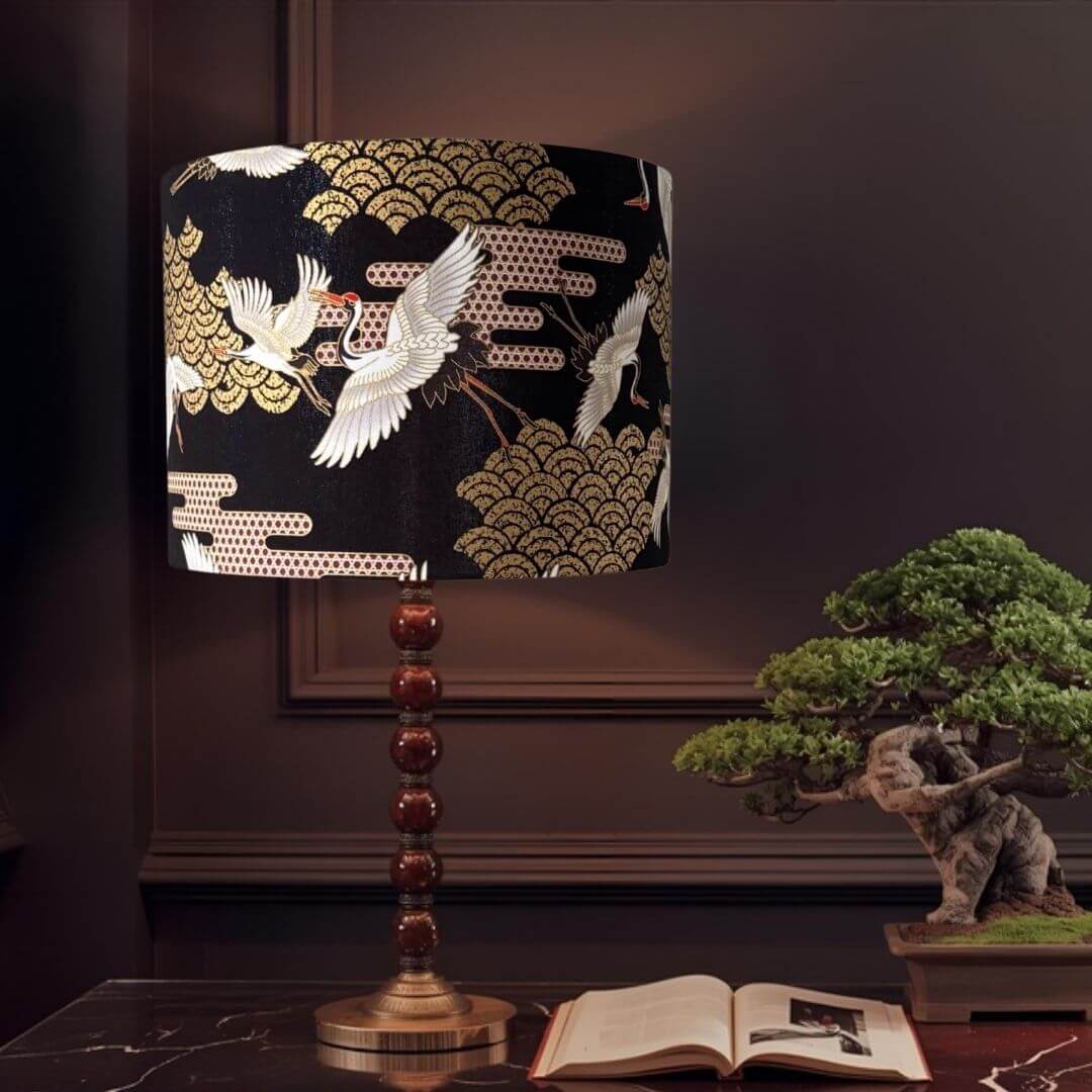 Japanese Cranes Lampshade, with cranes in flight and Art Deco-style clouds on a black background with gold metallic accents shown on a table with a bosai tree